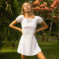 Woman in a short-sleeve white eyelet dress standing in a garden with greenery and flowers.