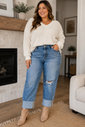 A woman modeling a cream ribbed knit sweater and distressed blue cuffed jeans with tan ankle boots, standing in a living room setting.