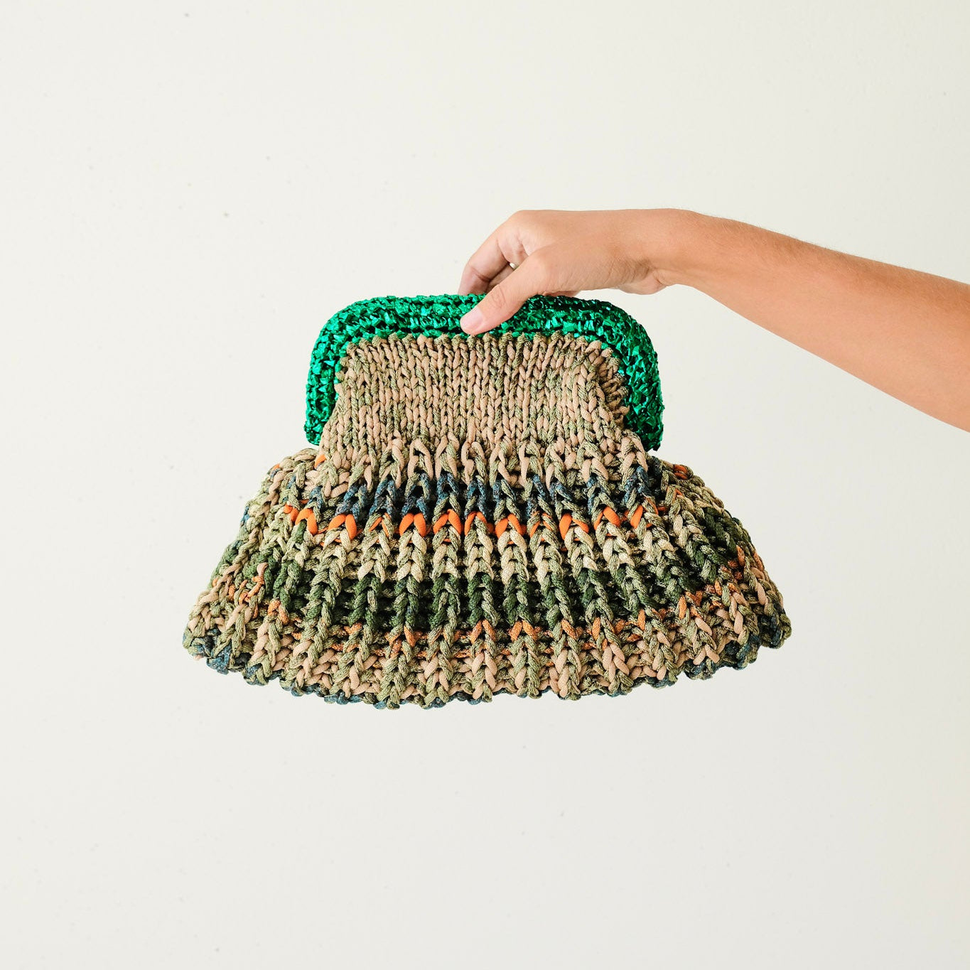 Hand holding a woven clutch bag with a green handle on a light background