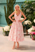 A woman wears the Florence Pink Floral Casual Midi Dress and sandals as she stands indoors, surrounded by potted plants and flowers.