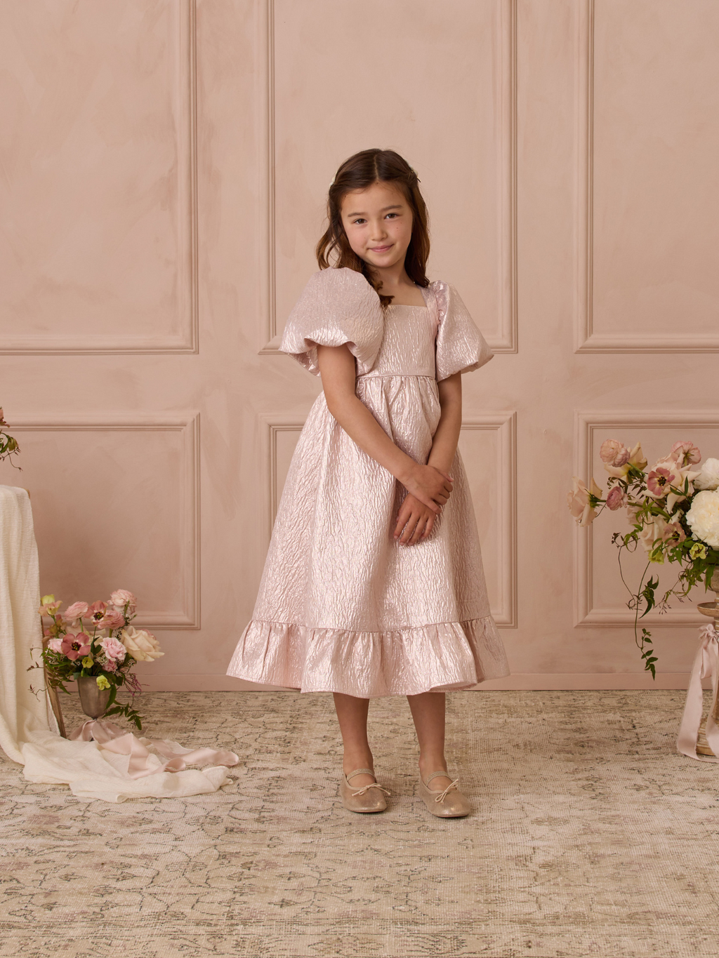 Young girl in a pink dress with puff sleeves, smiling in a softly lit room with floral decorations.