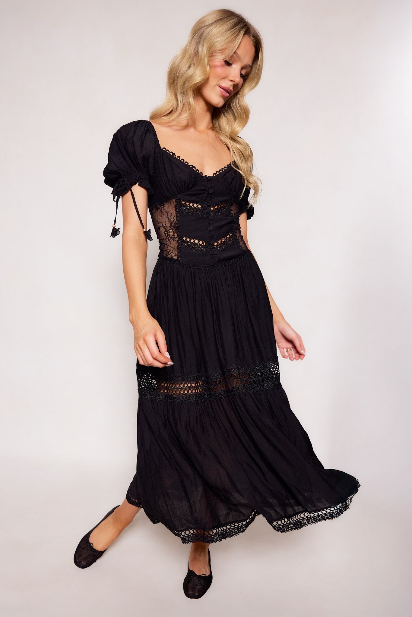 A woman with long blonde hair wears the Peregrine Dress in Black by Ivy City Co, featuring lace details and puffed sleeves. She stands against a plain light background, smiling softly while looking down.