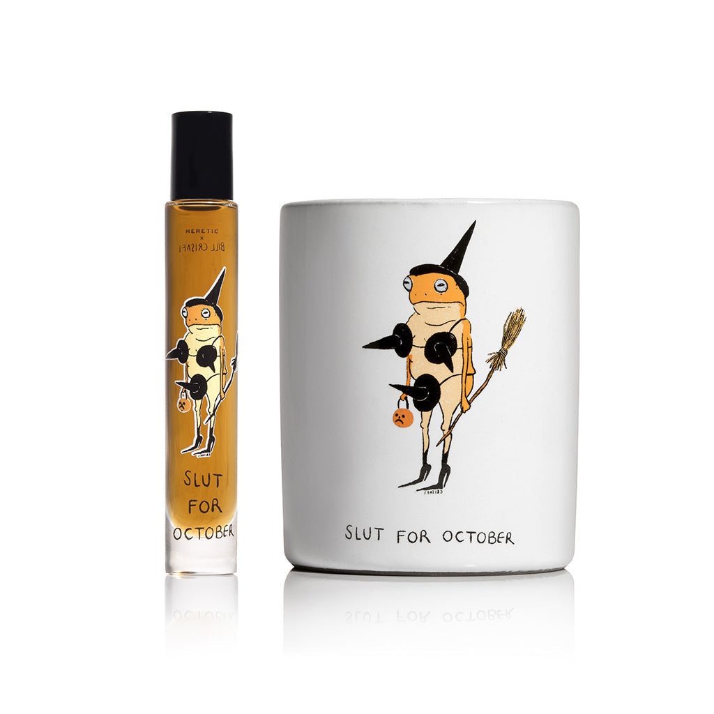 A small clear perfume bottle with amber liquid and a black cap stands beside a candle printed with a cartoon frog dressed as a witch holding a broom and pumpkin.