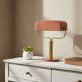 Modern table lamp with a pink shade and gold base on a white surface with a plant and book.